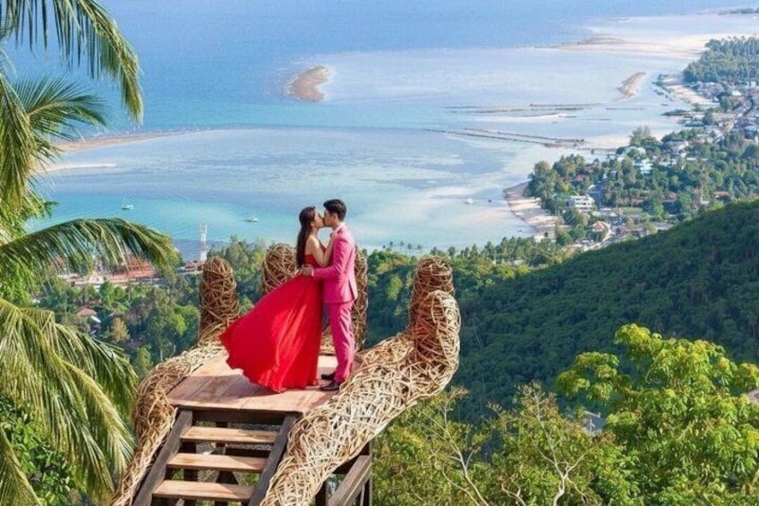 A couple on a wooden platform overlooking a coastal landscape with a beach and forested hills. 1