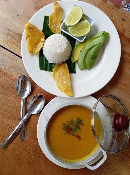 A plate with rice, avocado slices, fried plantains, lime wedges, and a bowl of soup garnished with herbs. 2