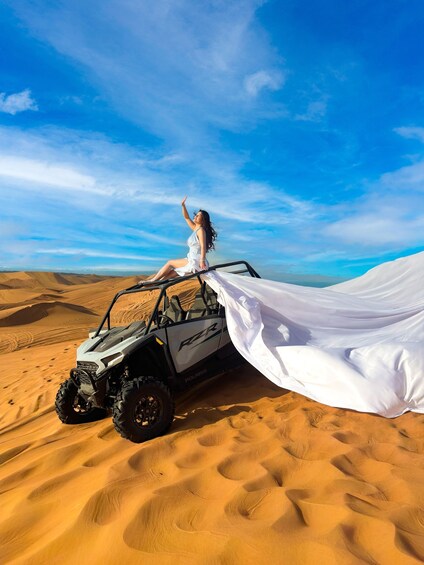 A person in a white dress sitting on an off-road vehicle in a desert with sand dunes and a clear sky. 1