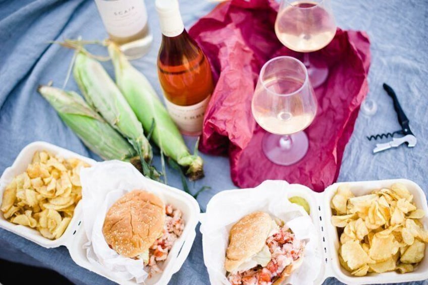 A picnic setup with food containers, corn on the cob, wine bottles, glasses, and a corkscrew. 2