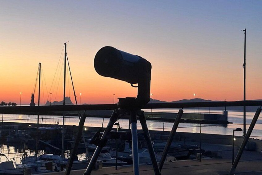 A telescope on a tripod overlooking a marina with boats and a bridge in the background during sunset. 1