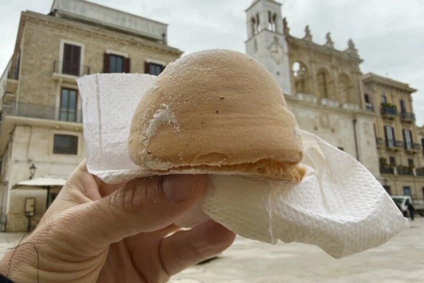 Une main tenant un sandwich avec une serviette en papier, devant un bâtiment historique doté d’un clocher. 2
