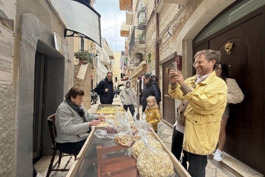 Une rue étroite où l’on voit des gens qui se promènent, un homme qui prend une photo et une table sur laquelle sont disposés des aliments. 3