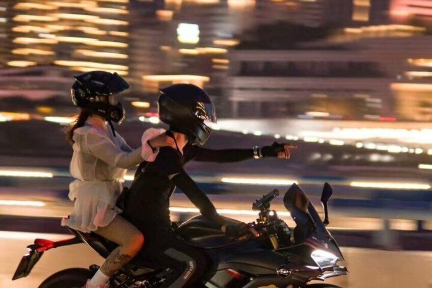 A person on a motorcycle with a passenger, both wearing helmets, in an urban setting with blurred lights in the background. 2