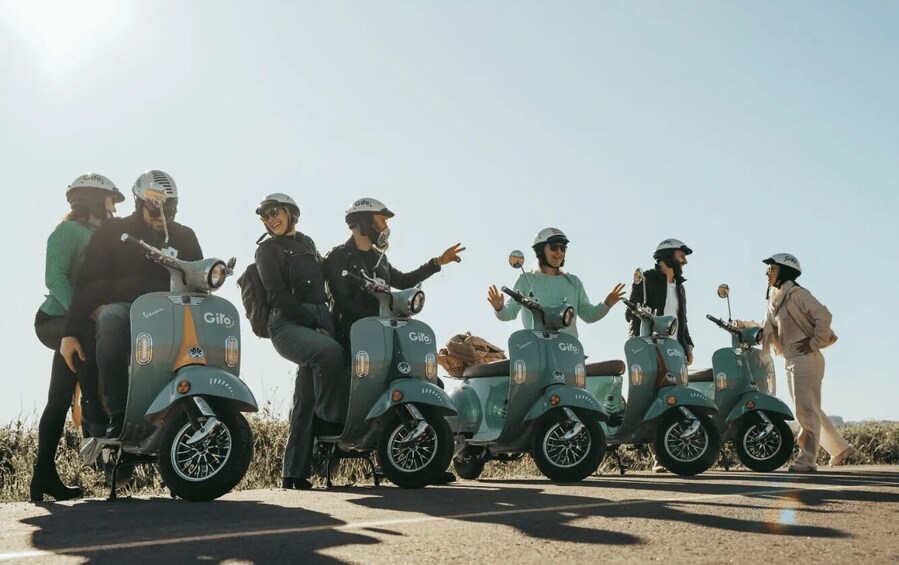 A group of people on scooters, some wearing helmets, on a road with a clear sky. 1