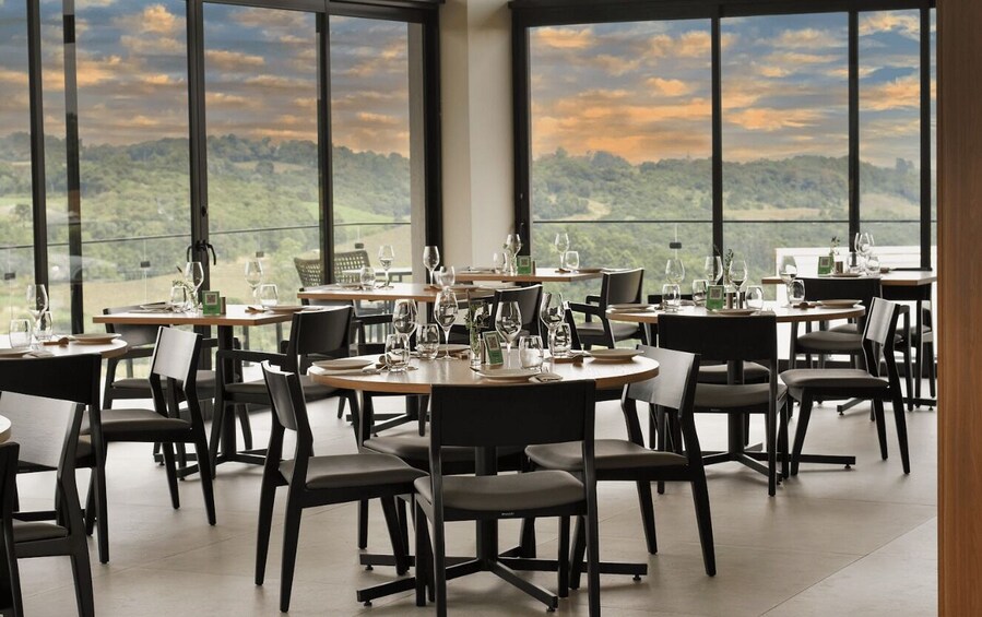 A restaurant with tables set for dining, large windows with a view of a hilly landscape. 2