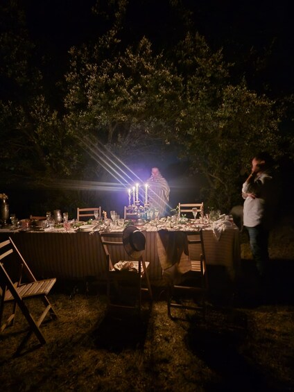 An outdoor dining setup at night with a table set for multiple guests, surrounded by trees. 2