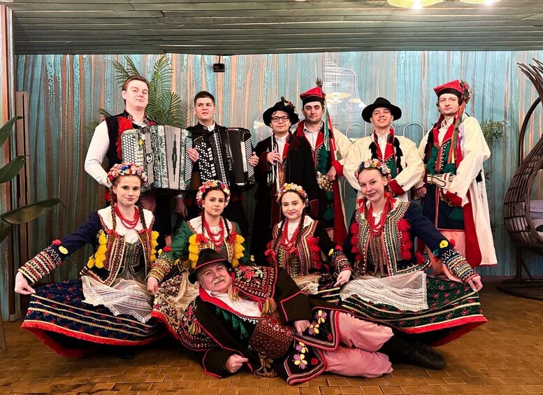A group of people in traditional costumes with musical instruments, seated and standing in a room with a rustic backdrop. 1