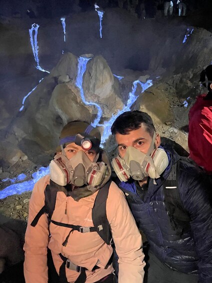 Two people wearing gas masks in a cave with glowing blue water. 2