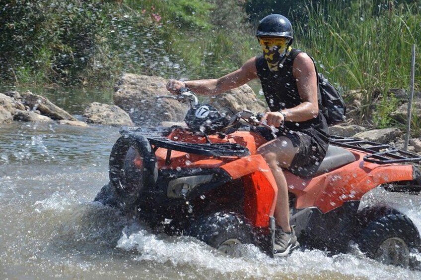 A person riding an ATV through a shallow water body surrounded by rocks and greenery. 3
