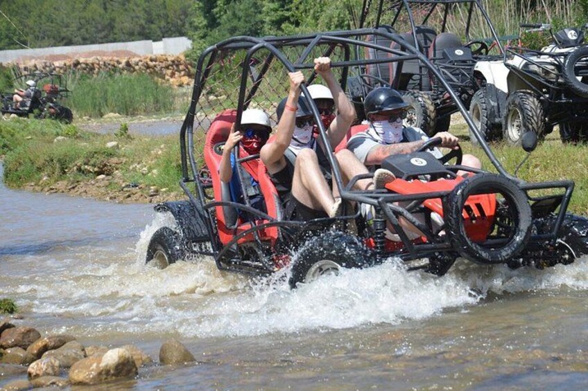 Two people in a red and black off-road vehicle navigating through a shallow river. 1