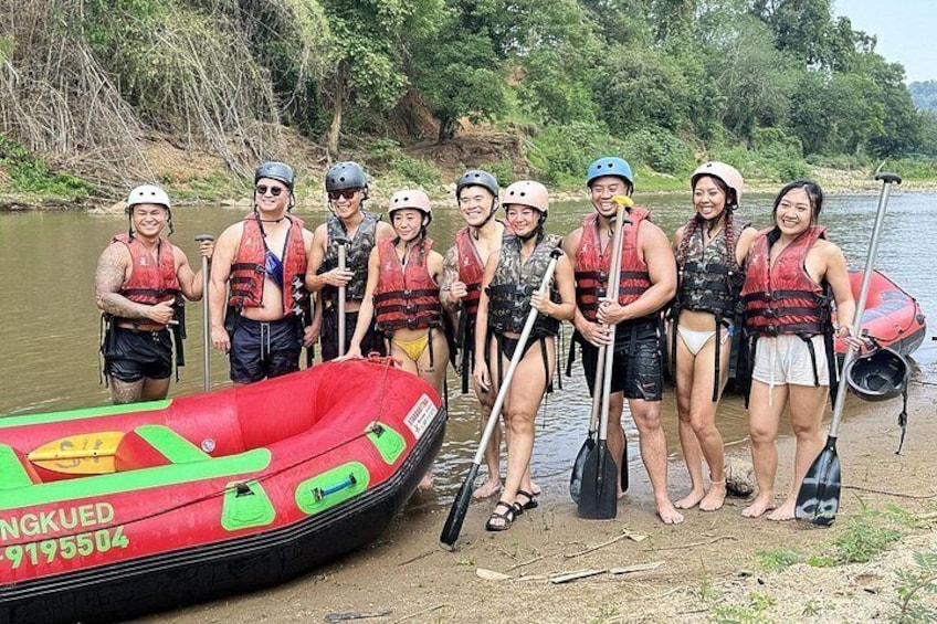 A group of people in life jackets and helmets by a river, with a red inflatable raft and paddles. 2