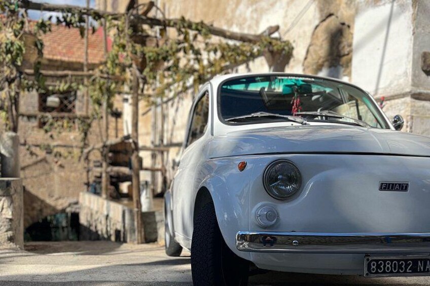 Un'auto Fiat d'epoca parcheggiata su una strada, con un muro in pietra e vegetazione sullo sfondo. 2