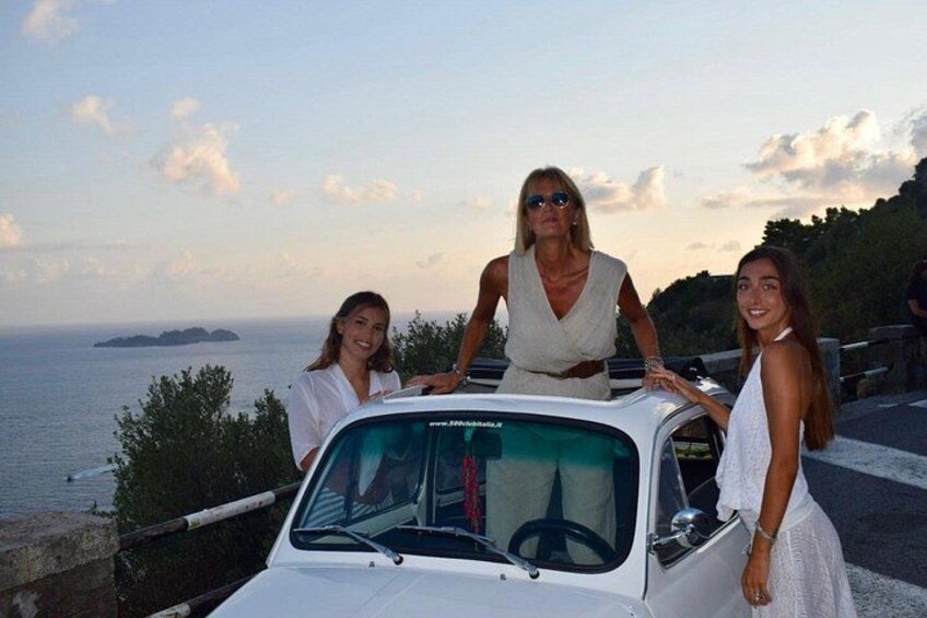 Tre persone in piedi accanto a un'auto bianca con vista sul mare e un paesaggio collinare sullo sfondo. 4