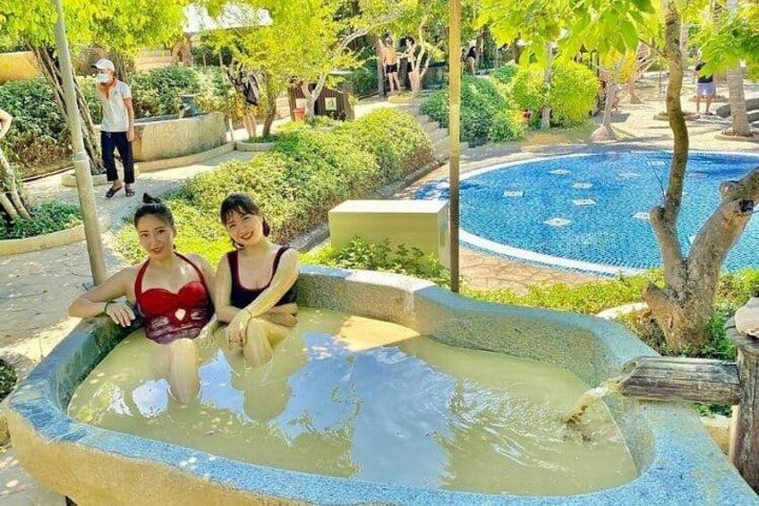 Twee personen zitten in een jacuzzi, omringd door groen en een zwembad. 2