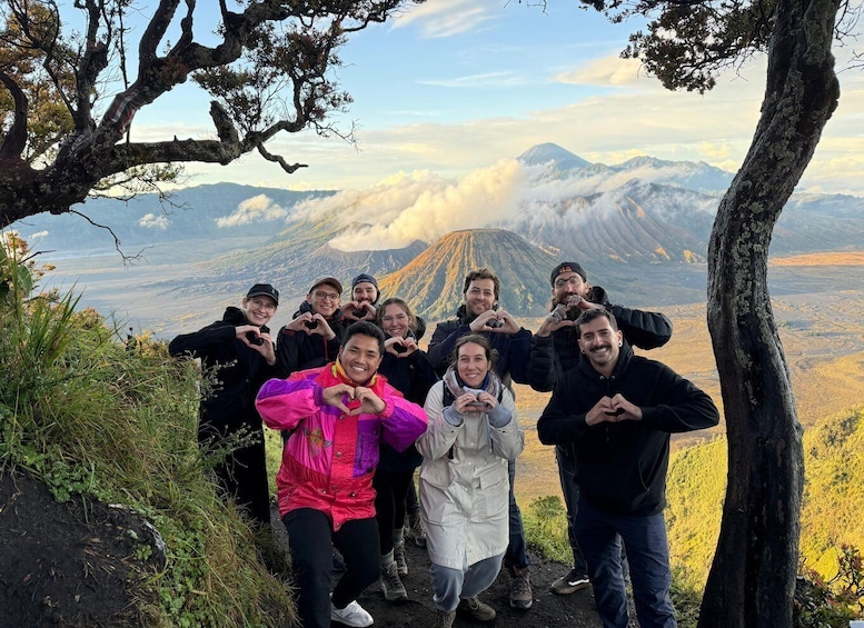 A group of people making heart shapes in front of a mountain with a forested area and a clear sky. 1