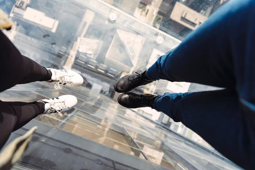 Deux paires de jambes portant des chaussures différentes, debout sur un sol de verre, avec une vue sur la ville en arrière-plan. 3