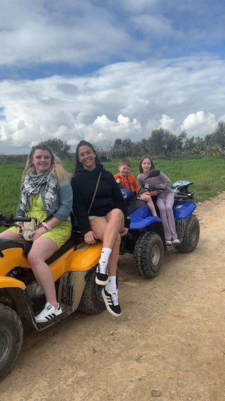 Four people on all-terrain vehicles on a dirt path with a grassy field and trees in the background. 3