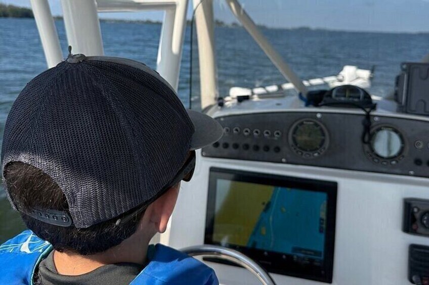 Eine Person mit Kopfbedeckung steuert ein Boot, das eine Bedienkonsole und einen Bildschirm mit einer Karte hat. 2