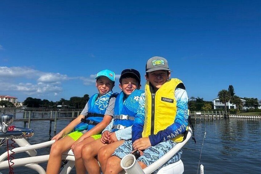 Drie mensen in een boot, met reddingsvesten, met kalm water en een steiger op de achtergrond. 5