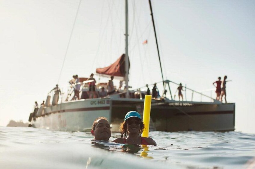 Twee mensen zwemmen in de oceaan, met een zeilboot op de achtergrond. 1