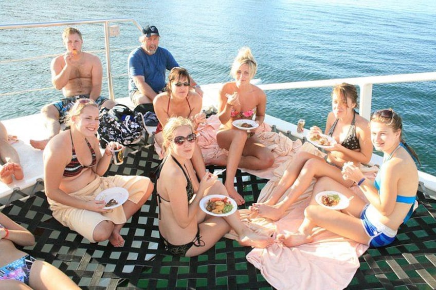 Een groep mensen op een boot, sommigen met borden eten in de hand, met water op de achtergrond. 2