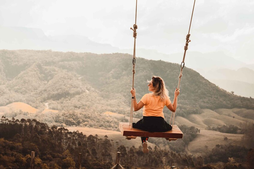 A person sitting on a swing suspended by ropes, overlooking a hilly landscape. 2