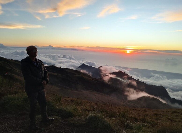 A person standing on a mountain ridge overlooking a valley with clouds below and a sunset in the distance. 1