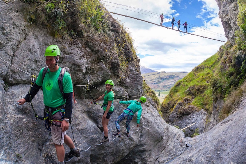 People rappelling down a rocky cliff, a rope bridge above, and a valley in the background. 3