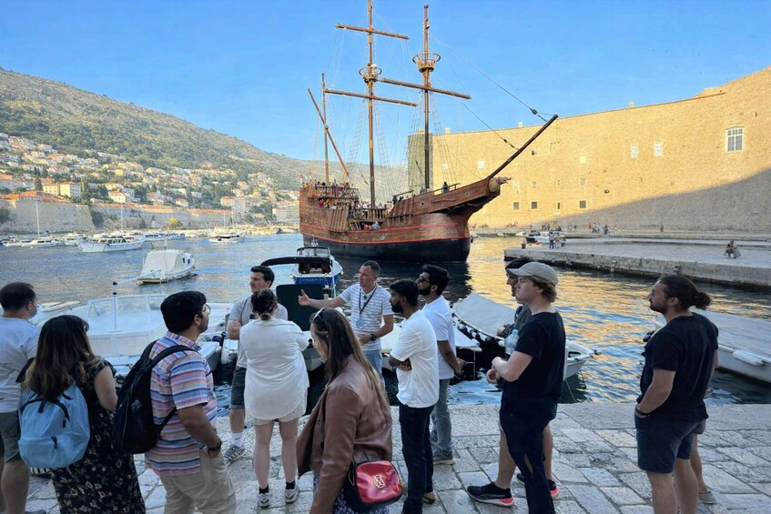 A group of people gathered around a large wooden ship docked at a harbor with a historic building in the background. 4