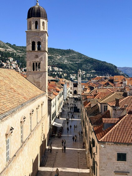 A historic town square with a prominent bell tower, terracotta-roofed buildings, and a hillside in the background. 2