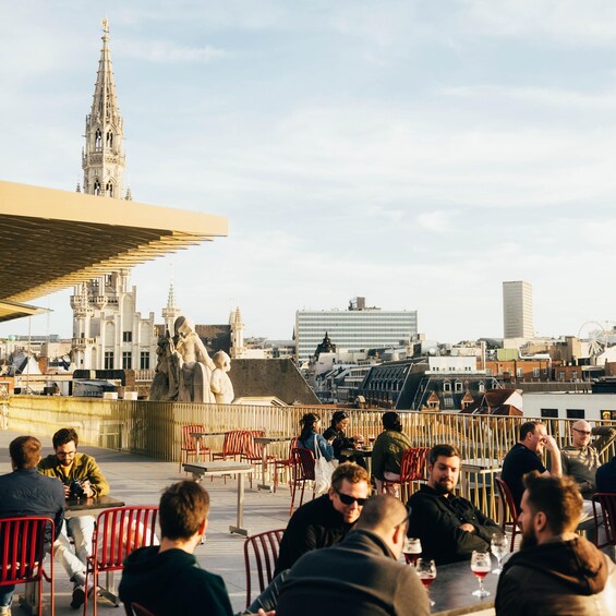 A rooftop bar with patrons seated at tables, overlooking a cityscape with historic architecture and a statue. 2