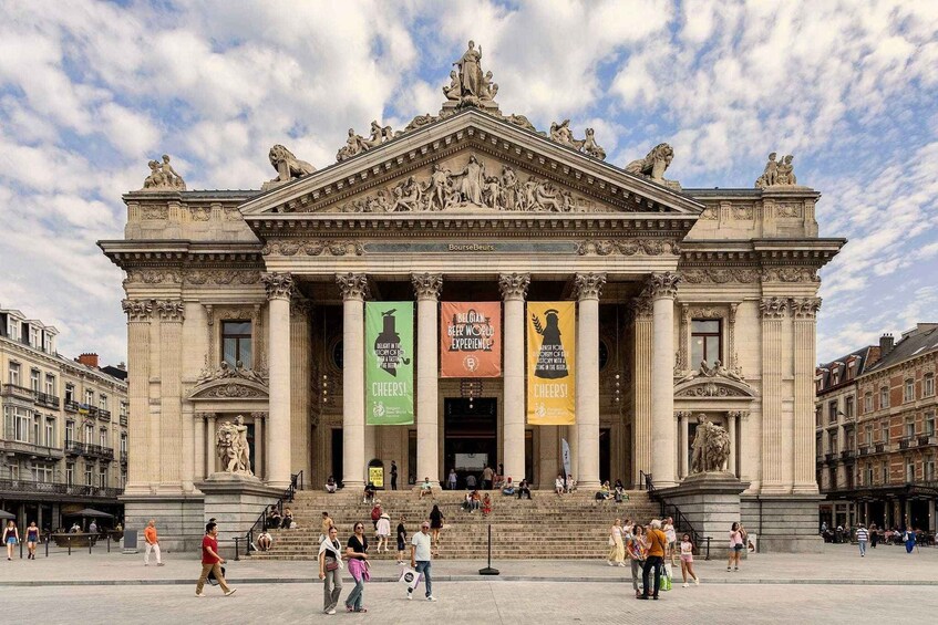 A grand neoclassical building with ornate sculptures and columns, banners hanging from the entrance, and people walking in front. 1