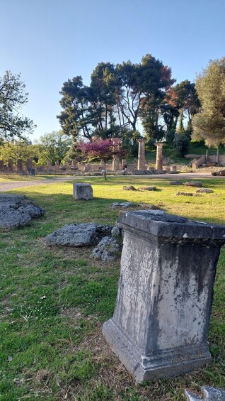 Ancient ruins with stone columns and a large stone slab with inscriptions, surrounded by grass and trees. 2