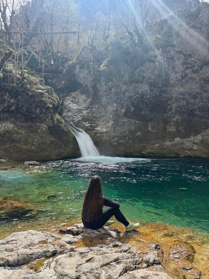 A person sitting on rocks by a clear, turquoise river with a waterfall in the background. 3