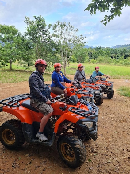 Four people on orange quad bikes in a rural setting with trees and a clear sky. 5