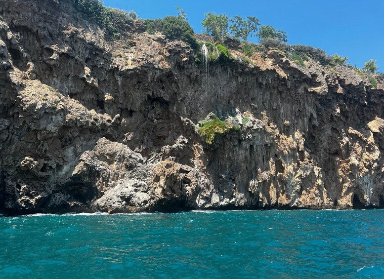 A rocky coastline with clear blue waters, a cliff with sparse vegetation, and a visible waterfall. 3