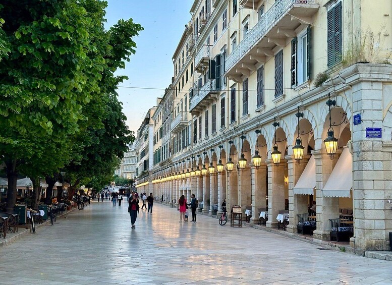 A tree-lined street with historic buildings, outdoor seating, and people walking. 1