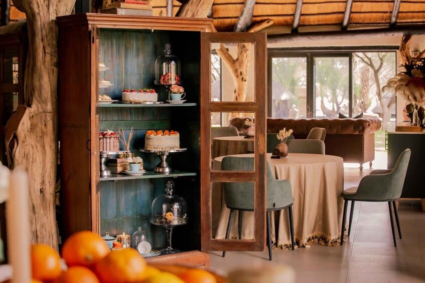 A rustic dining area with a wooden cabinet displaying cakes and pastries, a round table with chairs, and a large window with a view of trees. 2
