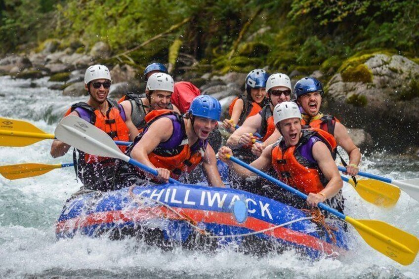 A group of people in a blue and red inflatable raft with 'CHILLIWACK' written on it, paddling through a river. 1