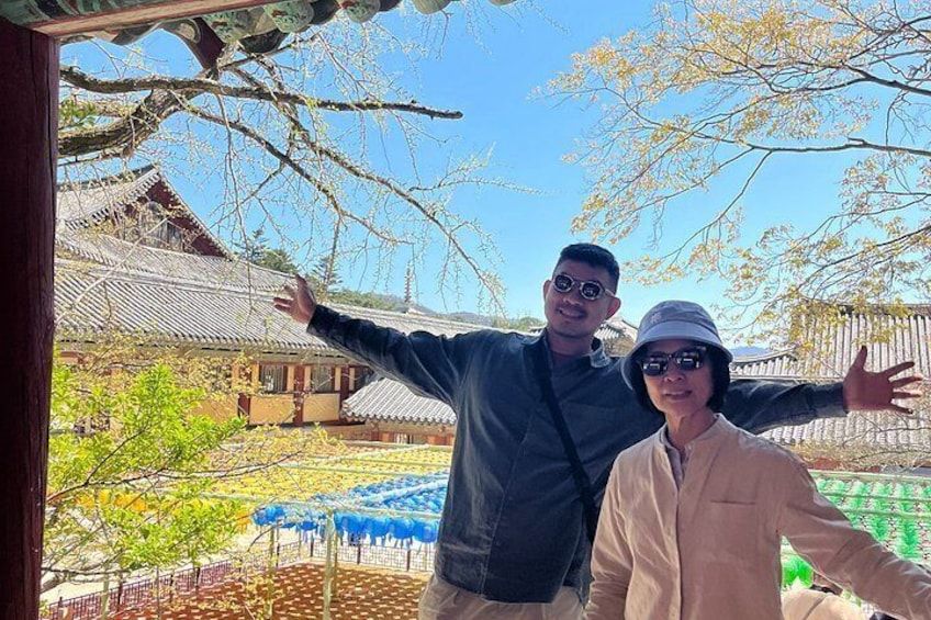 Two people standing in front of a traditional building with a tiled roof, a tree with sparse branches, and a fenced area with blue umbrellas. 3