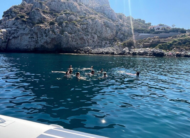 People swimming in a clear blue sea with rocky cliffs and a building in the background. 2