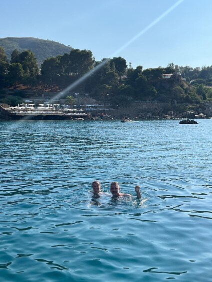 Three people swimming in a clear blue sea with a coastal town and hills in the background. 5