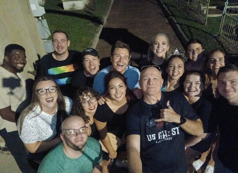 A group of people outdoors at night, some wearing casual clothing, with a building and a fence in the background. 3
