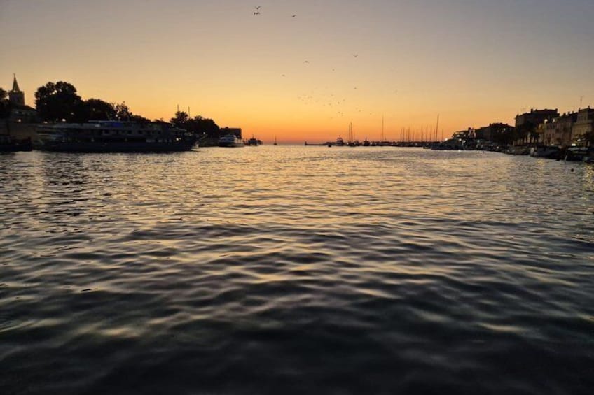Un coucher de soleil sur une rivière paisible, avec des bateaux, des bâtiments et le profil d’une ville lointaine à l’horizon. 3