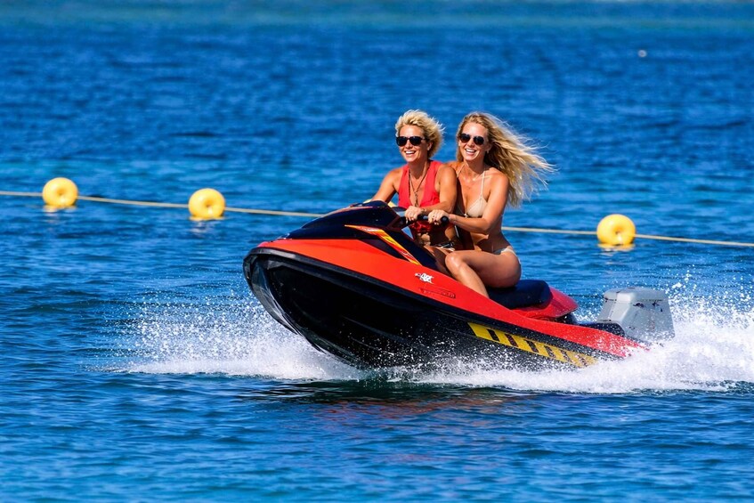 Two people on a red and black jet ski on a body of water with buoys. 1