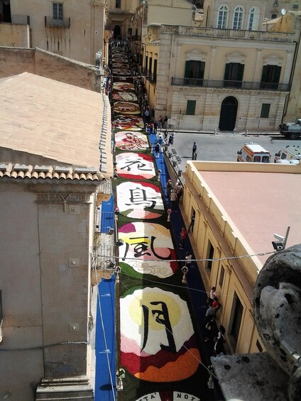 A street with a colorful, patterned carpet laid out along the sidewalk, bordered by buildings. 5