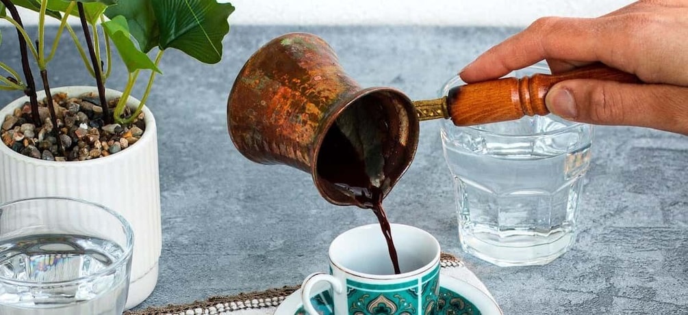 A hand pouring a dark liquid from a copper cup into a white mug. 3
