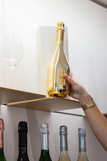 A hand holding a champagne bottle with a gold foil top, placed on a shelf with other bottles and a wine glass. 1