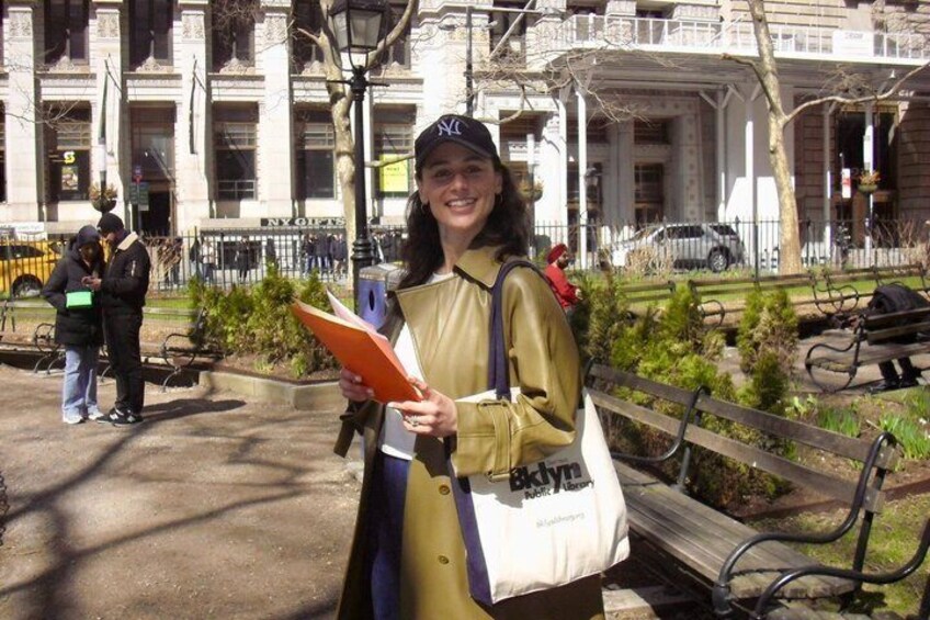 A person holding a book and a bag, standing in a park with benches and a building in the background. 1